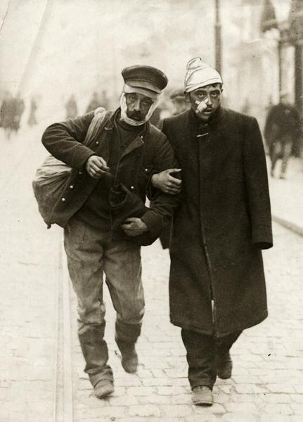 A wounded German soldier and a wounded Belgian soldier walk arm in arm down a street, 1915.