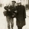 A wounded German soldier and a wounded Belgian soldier walk arm in arm down a street, 1915.